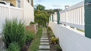 Un sendero estrecho bordeado por cercas blancas y abundante vegetación, que conduce a un edificio de fachada blanca y puerta verde.
