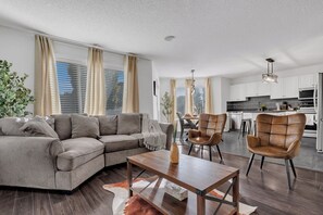 Een moderne woonkamer met een hoekbank, een salontafel en twee leren fauteuils. Achtergrond toont een keuken met witte kasten en roestvrijstalen apparatuur.