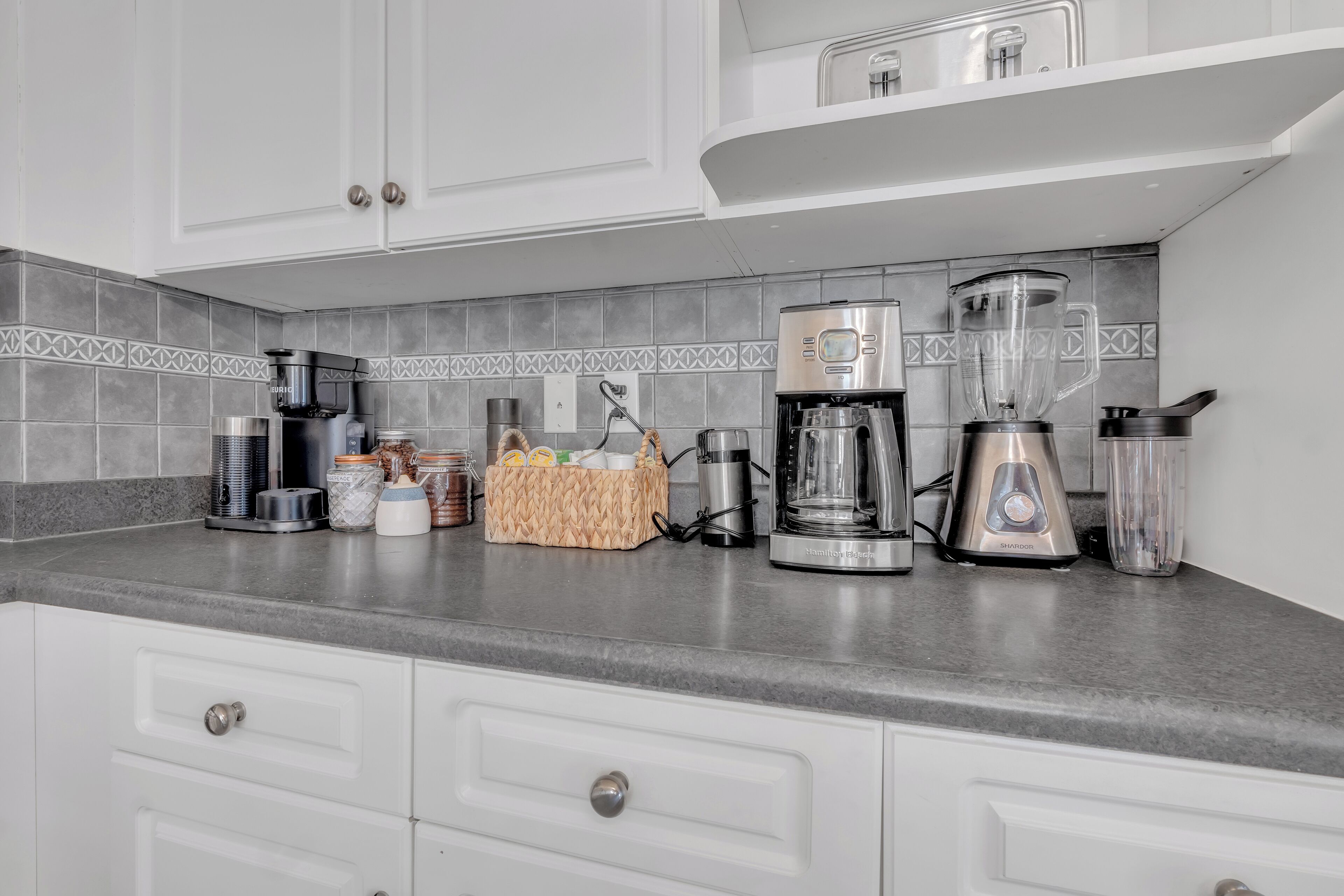 A kitchen with white cabinets, a coffee maker, a blender, and a basket of items on the counter.