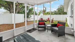 Un patio couvert aménagé avec des meubles en osier, offrant une vue sur le jardin et bordé d’une clôture blanche.