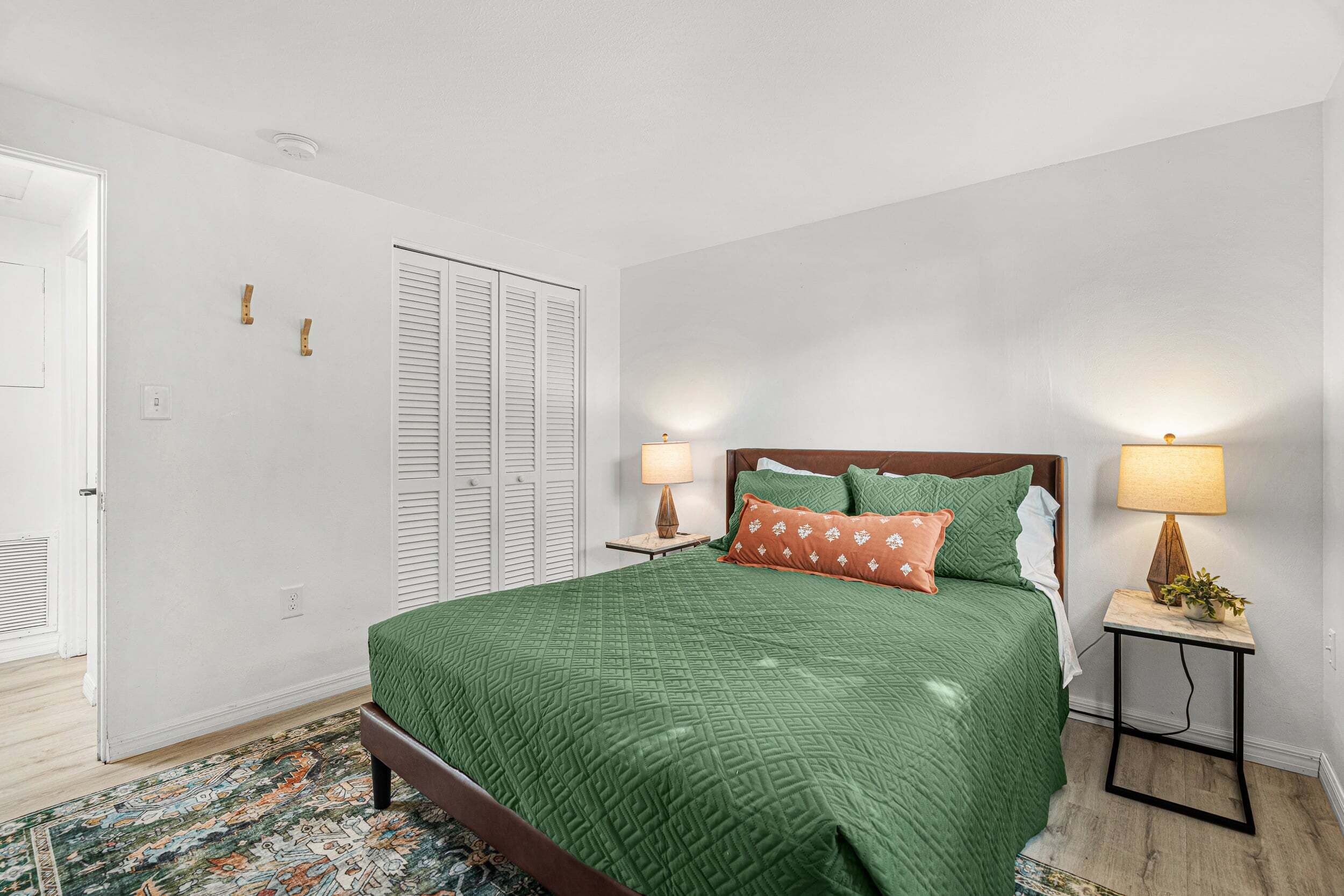 A bedroom with a bed, bedside lamps, a nightstand, a patterned rug, and a door with a closet.