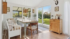 Una stanza con soffitto in legno bianco, un tavolo da pranzo con sedie, un armadietto in legno e un'ampia finestra che dà su del verde all'esterno.