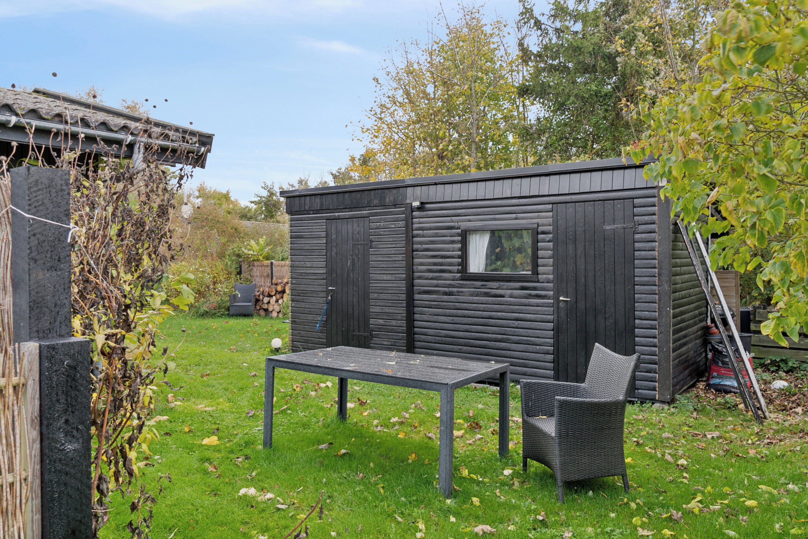 A small wooden cabin with a table and chair in front, surrounded by grass and trees.