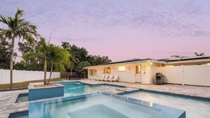 A modern outdoor swimming pool with a hot tub, surrounded by a white fence, palm trees, and a patio area with lounge chairs. - Olive Tree Oasis | 4/2 w/ Hot Tub + Heated Pool (Sarasota)