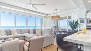 Ein modernes Wohnzimmer mit einer großen Eckcouch, einem Essbereich mit weißem Tisch und einer Küche mit großem Fenster mit Meerblick.
