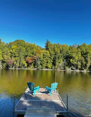 Une jetée en bois avec deux fauteuils Adirondack turquoise, placés face à un lac paisible, lui-même entouré d’une forêt dense.