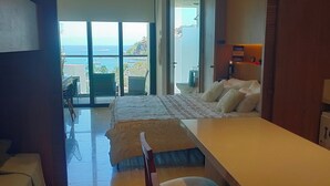 A modern hotel room with a large bed, a desk, and a balcony with a view of the sea.