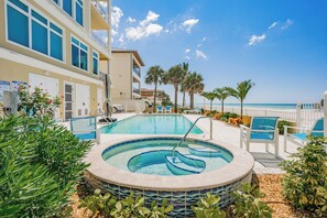 A swimming pool with a hot tub, surrounded by palm trees and beach chairs, with a view of the ocean. - Sea Star Penthouse—Luxury Beachfront Escape w/ Private Elevator, Gulf Views & Designer Touches (Indian Shores)
