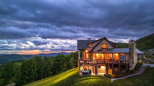A large, two-story house with a stone chimney, surrounded by lush greenery and a mountainous backdrop. - Over The Top: Custom Luxury Home! Views, Hot Tub, Pool Table, Firepit, Claw Machine, More! (Banner Elk)