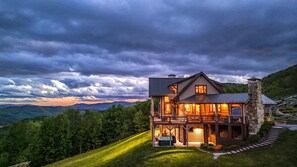 A large, two-story house with a stone chimney, surrounded by lush greenery and a mountainous backdrop. - Over The Top: Custom Luxury Home! Views, Hot Tub, Pool Table, Firepit, Claw Machine, Sleeps 23! (Banner Elk)