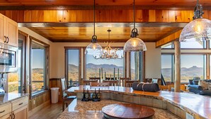 A spacious kitchen with granite countertops, wooden cabinets, and a large island. There are two pendant lights with a unique chandelier design above the island. - Over The Top: Custom Luxury Home! Views, Hot Tub, Pool Table, Firepit, Claw Machine, More! (Banner Elk)