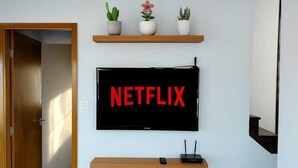 A modern living room with a mounted TV displaying the Netflix logo, a wooden shelf with potted plants, and a staircase leading to another level. - Best Location in Rio Vermelho, walking distance to the beach and fun (Salvador)