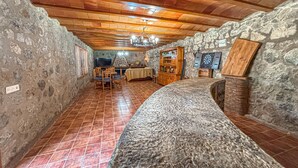 Una habitación rústica de paredes de piedra, techo de madera y suelo de baldosas, con zona de comedor con sillas y una mesa, un televisor y un dardo.