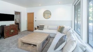 Ein modernes Wohnzimmer mit einem großen Fenster, einem Holztisch, einem weißen Sofa mit Kissen und einer roten, alten Kommode.
