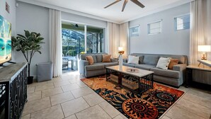 A modern living room with a grey sofa, a coffee table, and a patterned rug. There is a ceiling fan, a TV, and a plant. - The villa where dreams come true! (Davenport)