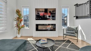 Un salon moderne avec un grand téléviseur à écran plat fixé au-dessus d’une cheminée, un confortable canapé gris, une petite table basse ronde sur laquelle sont posés des livres et une plante en pot.