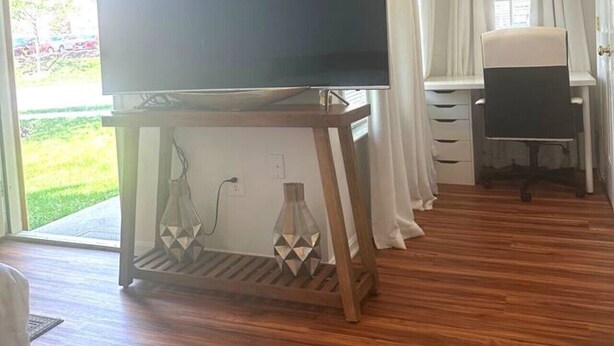 Ein Zimmer mit einem großen Flachbildfernseher auf einem Holzständer, einem Schreibtisch mit Stuhl und Blick ins Grüne.