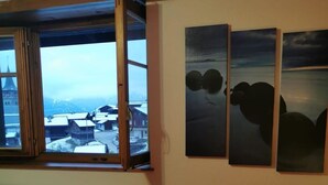 Une chambre avec un lit, une fenêtre donnant sur un village et trois tableaux accrochés au mur.