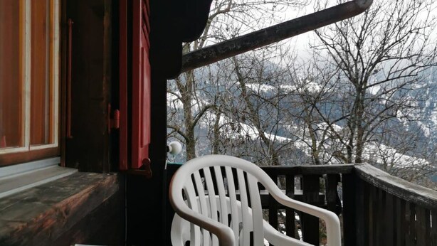 Une table recouverte d’une nappe à motifs floraux, une bougie allumée, une chaise en plastique blanche et une vue sur un paysage enneigé, visible à travers une rambarde en bois.