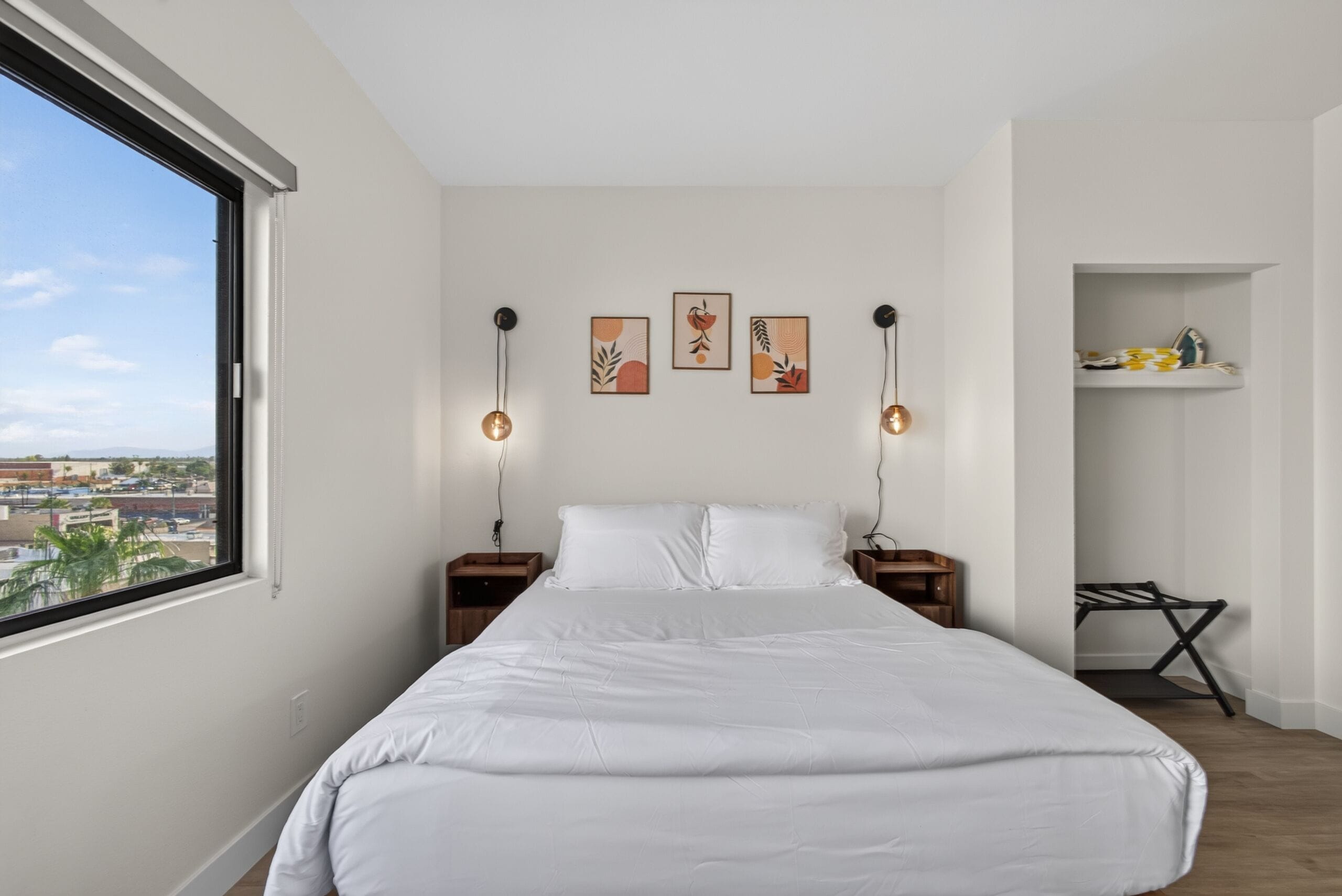 A bedroom with a large bed, two bedside tables, a wall-mounted shelf, and a window with a city view.