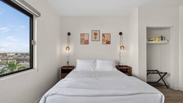 A bedroom with a large bed, two bedside tables, a wall-mounted shelf, and a window with a city view.