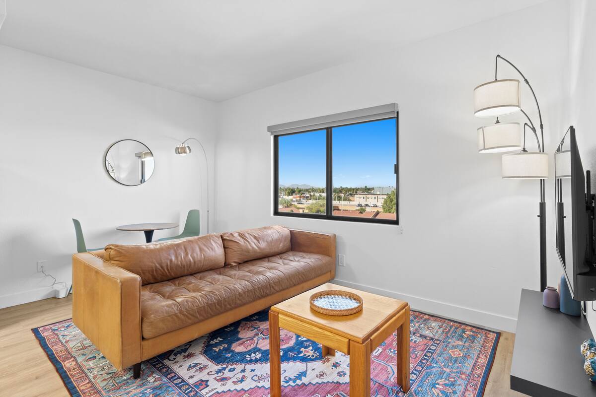 A modern living room with a leather sofa, a small wooden table, a patterned rug, a round mirror, and a large window offering a city view.