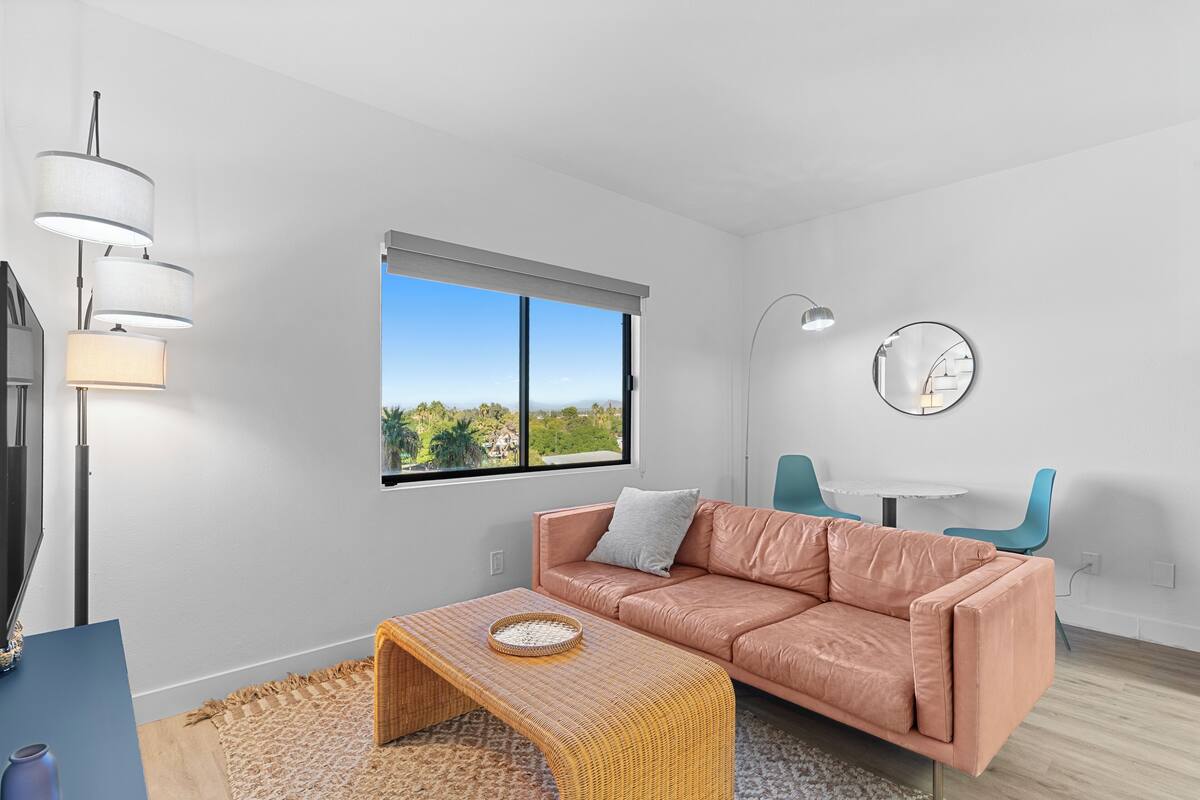 A modern living room with a leather sofa, a wicker coffee table, a flat-screen TV, and a large window offering a view of greenery.