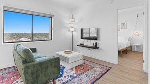 Una sala moderna con un ventanal grande, un sillón verde, una mesa de centro blanca, un televisor montado en la pared y una cama en la habitación contigua.