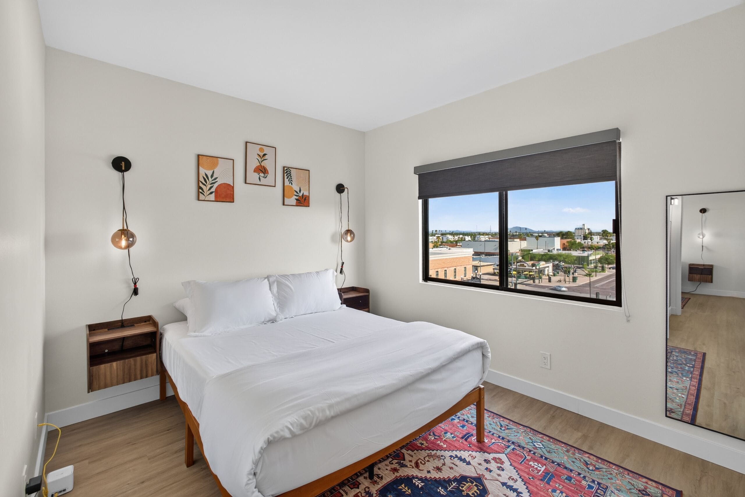 A bedroom with a bed, a nightstand, a wall-mounted lamp, a window with a view of buildings, and a mirror.