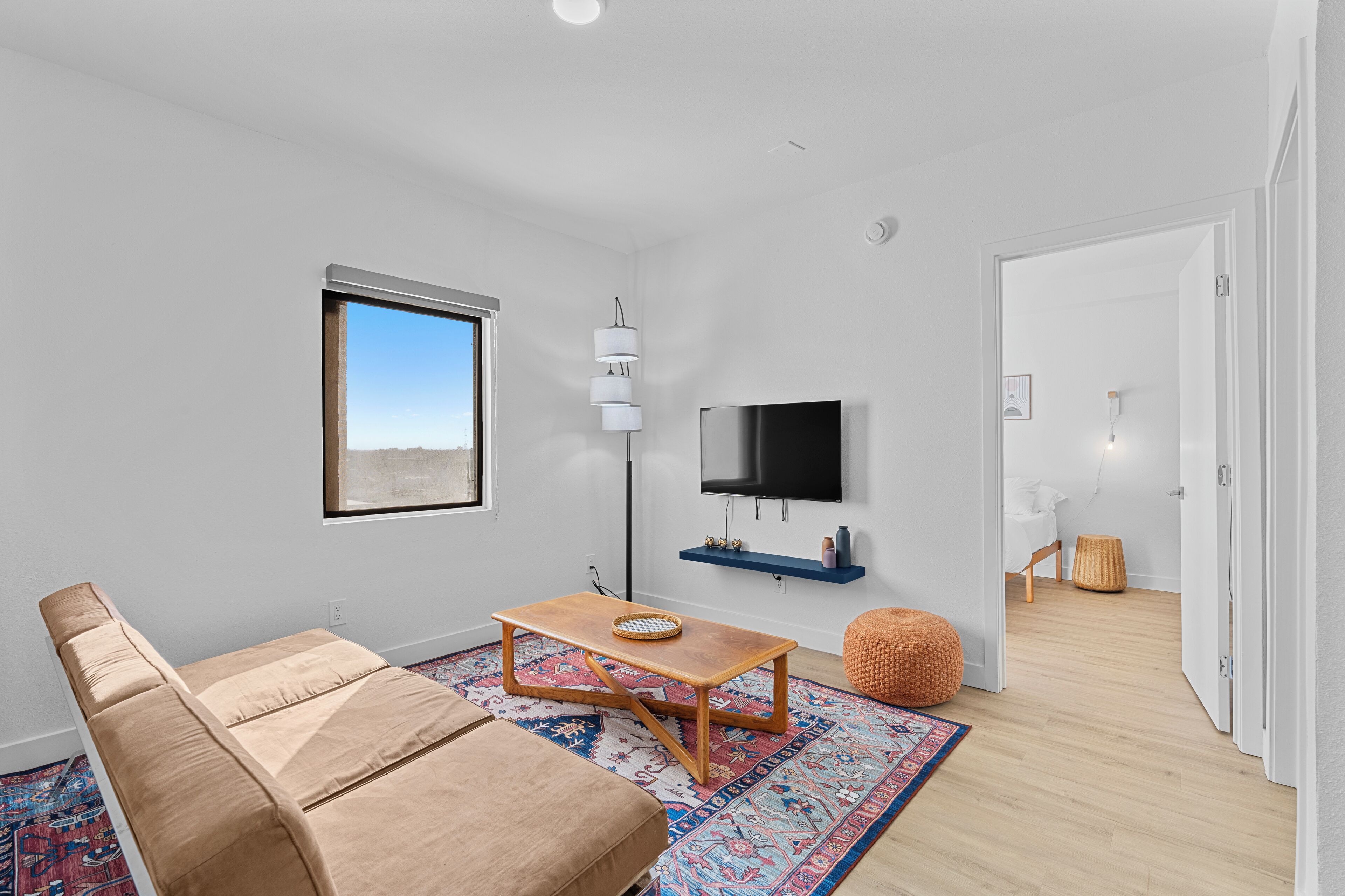 A modern living room with a sofa, a coffee table, a TV mounted on the wall, and a view of the outdoors through a window.