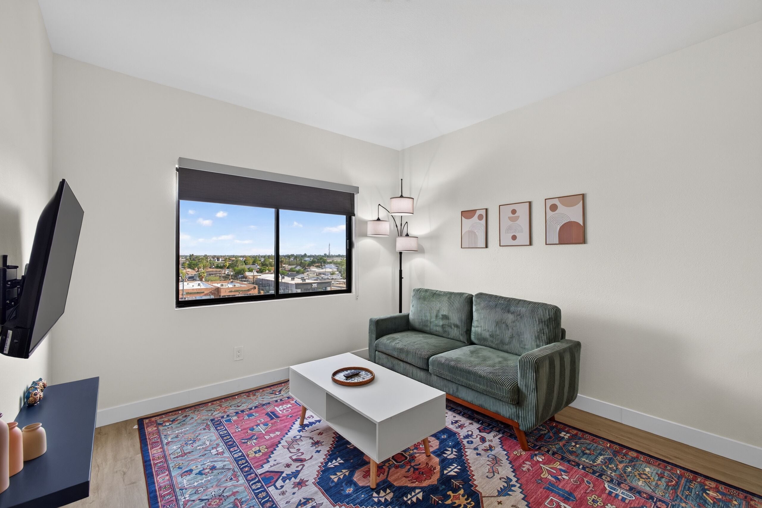 A modern living room with a green sofa, a white coffee table, a large window with a city view, and a floor-to-ceiling lamp.