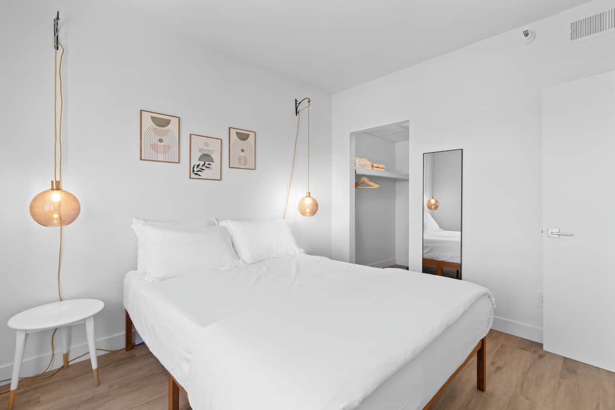 A modern bedroom with a large bed, two pendant lights, a small white stool, and a walk-in closet.