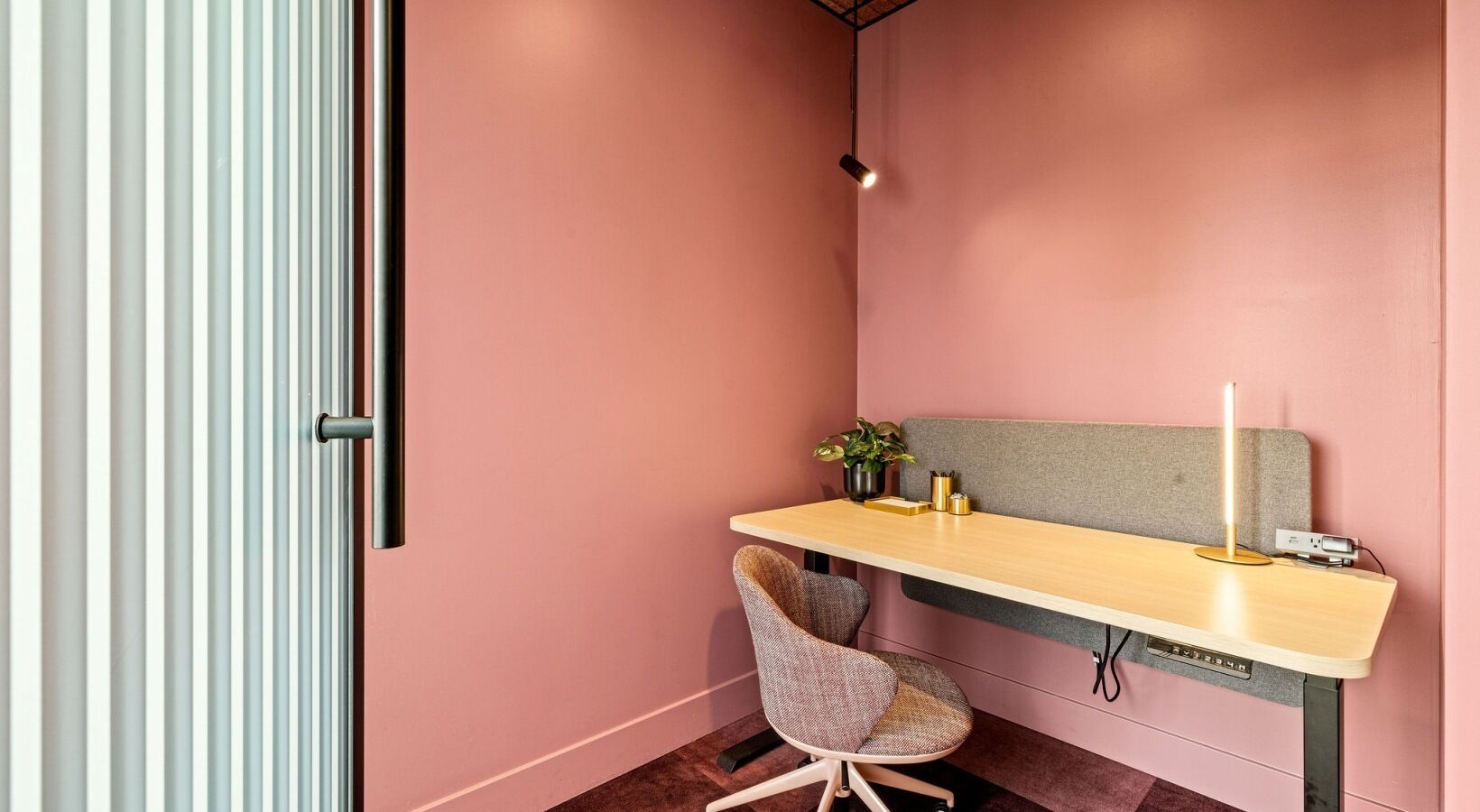 A small, pink-walled room with a wooden desk, a cushioned chair, a plant, and a candle.