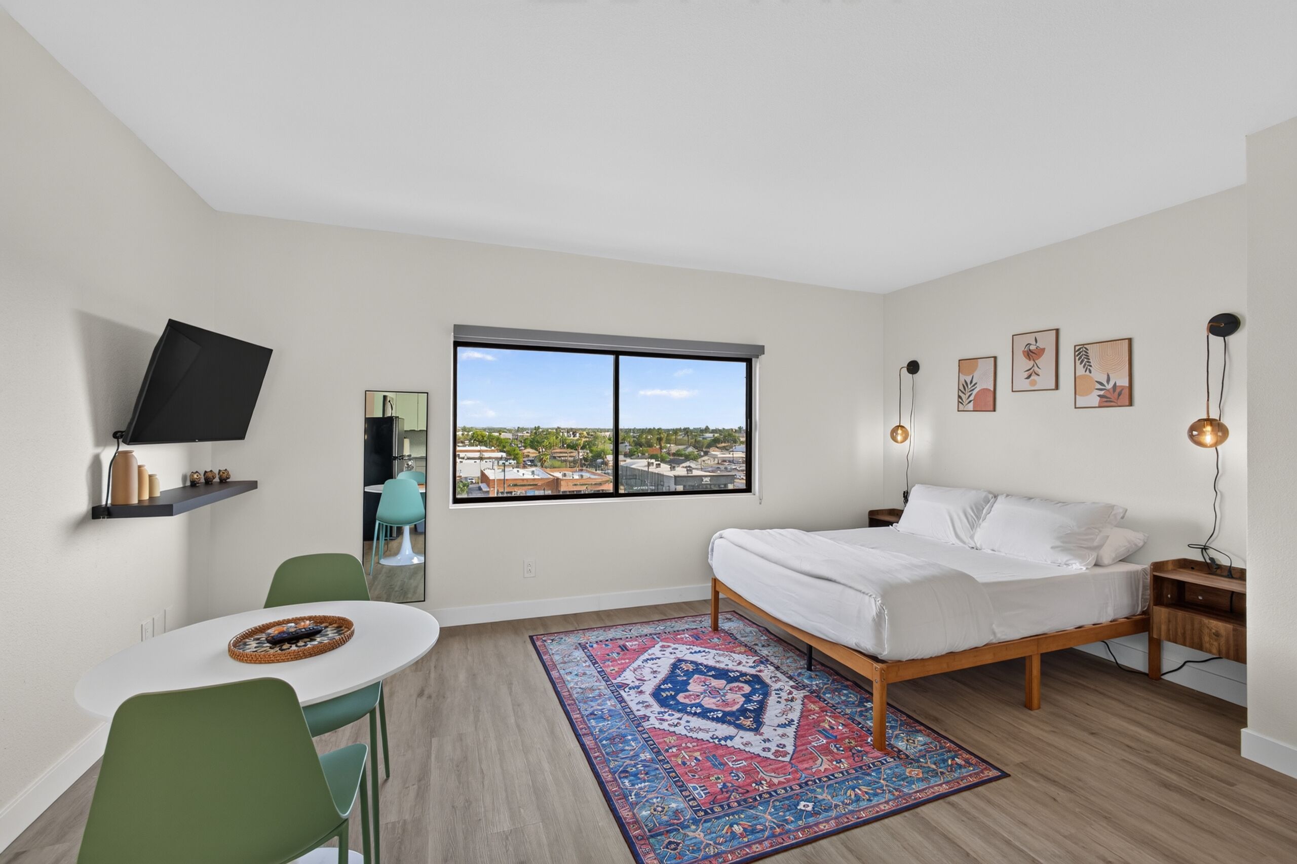 A modern bedroom with a bed, a desk, a chair, a large window with a city view, and a TV mounted on the wall.