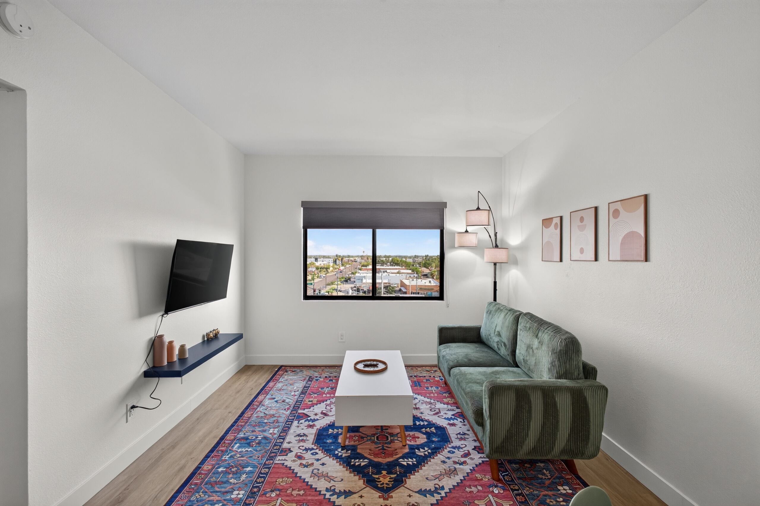 A modern living room with a flat-screen TV, a sofa, a coffee table, a patterned rug, and a window with a city view.