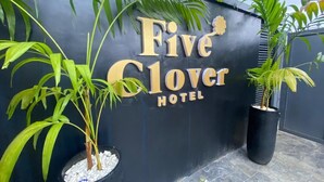 Et skilt for Five Clover Hotel med store gyldne bogstaver på en mørk væg, omkranset af potteplanter.