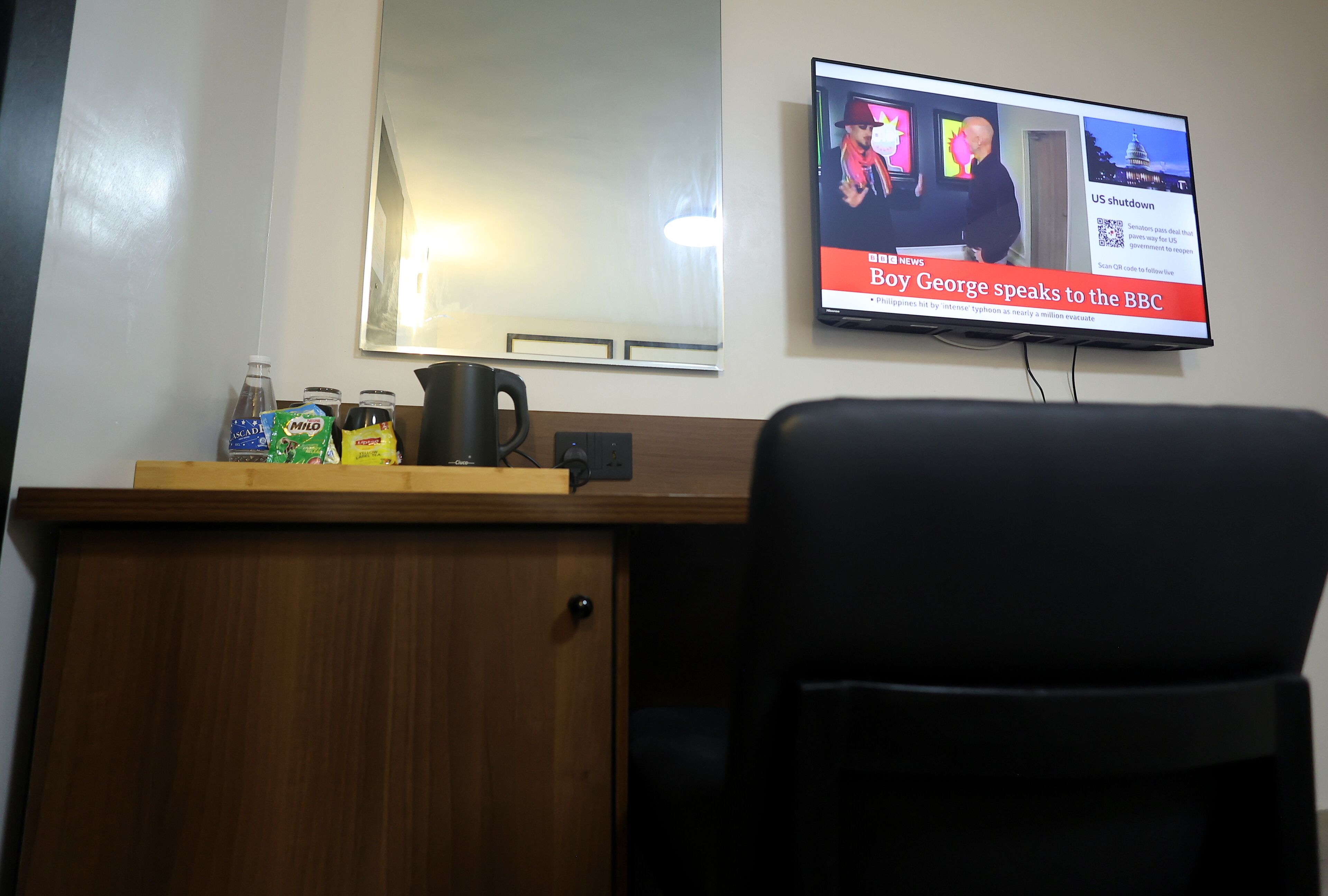 Une pièce avec un téléviseur diffusant un programme d’information, un bureau en bois avec une cafetière et une chaise.