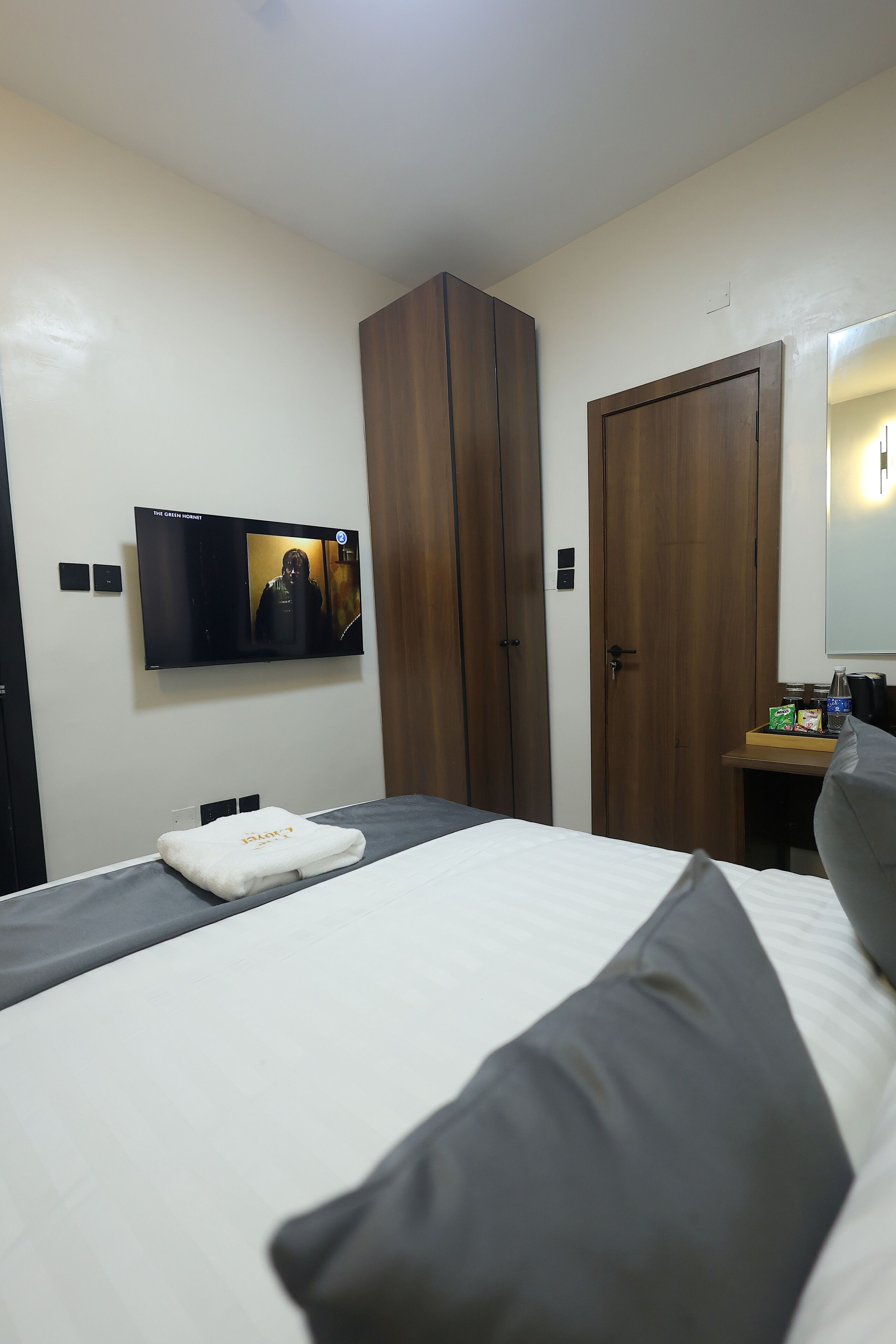 A hotel room with a bed, a TV mounted on the wall, a wardrobe, and a small table with a bottle and a box.