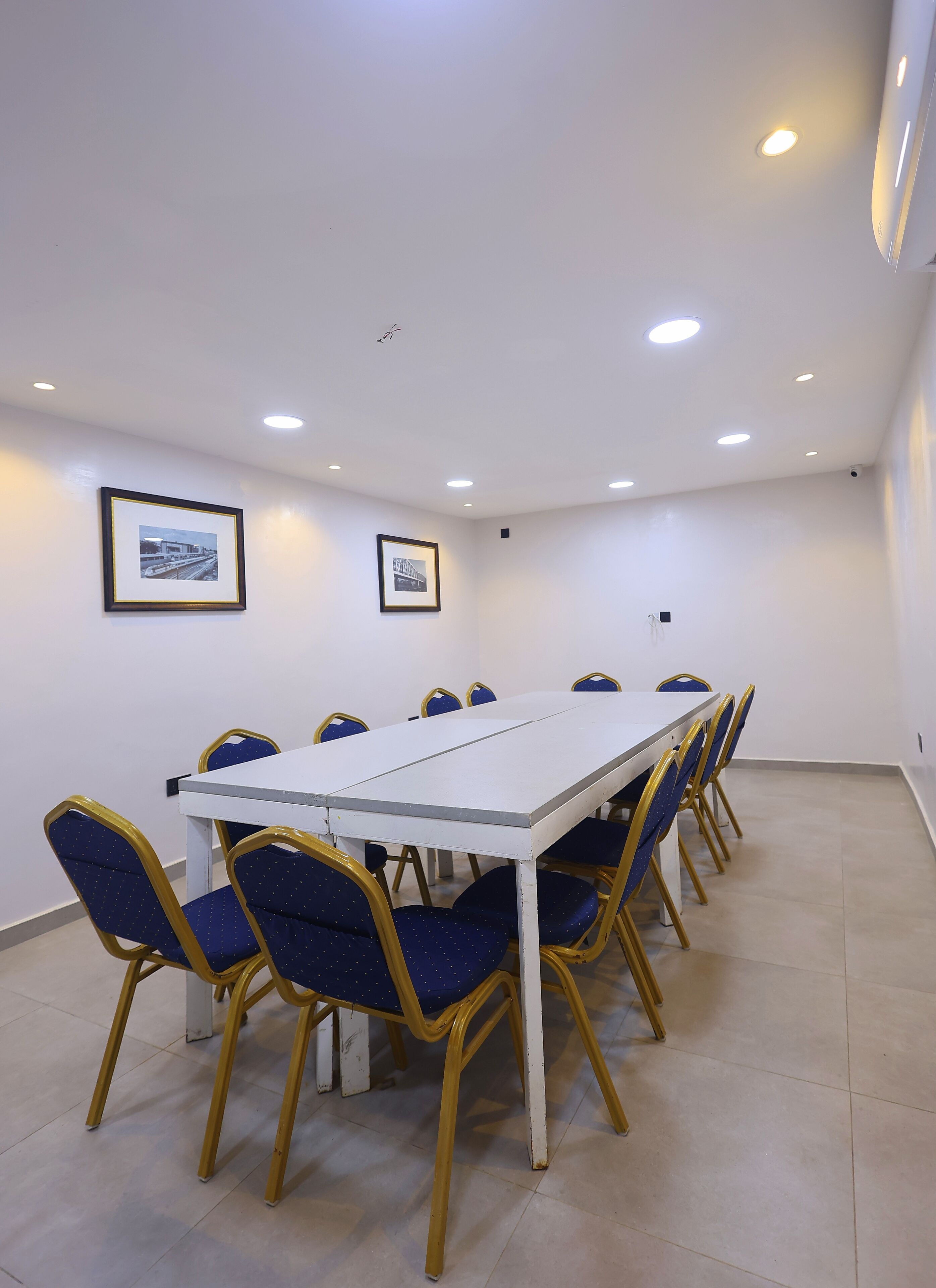 A conference room with a white table and blue chairs, framed pictures on the walls, and recessed ceiling lights.