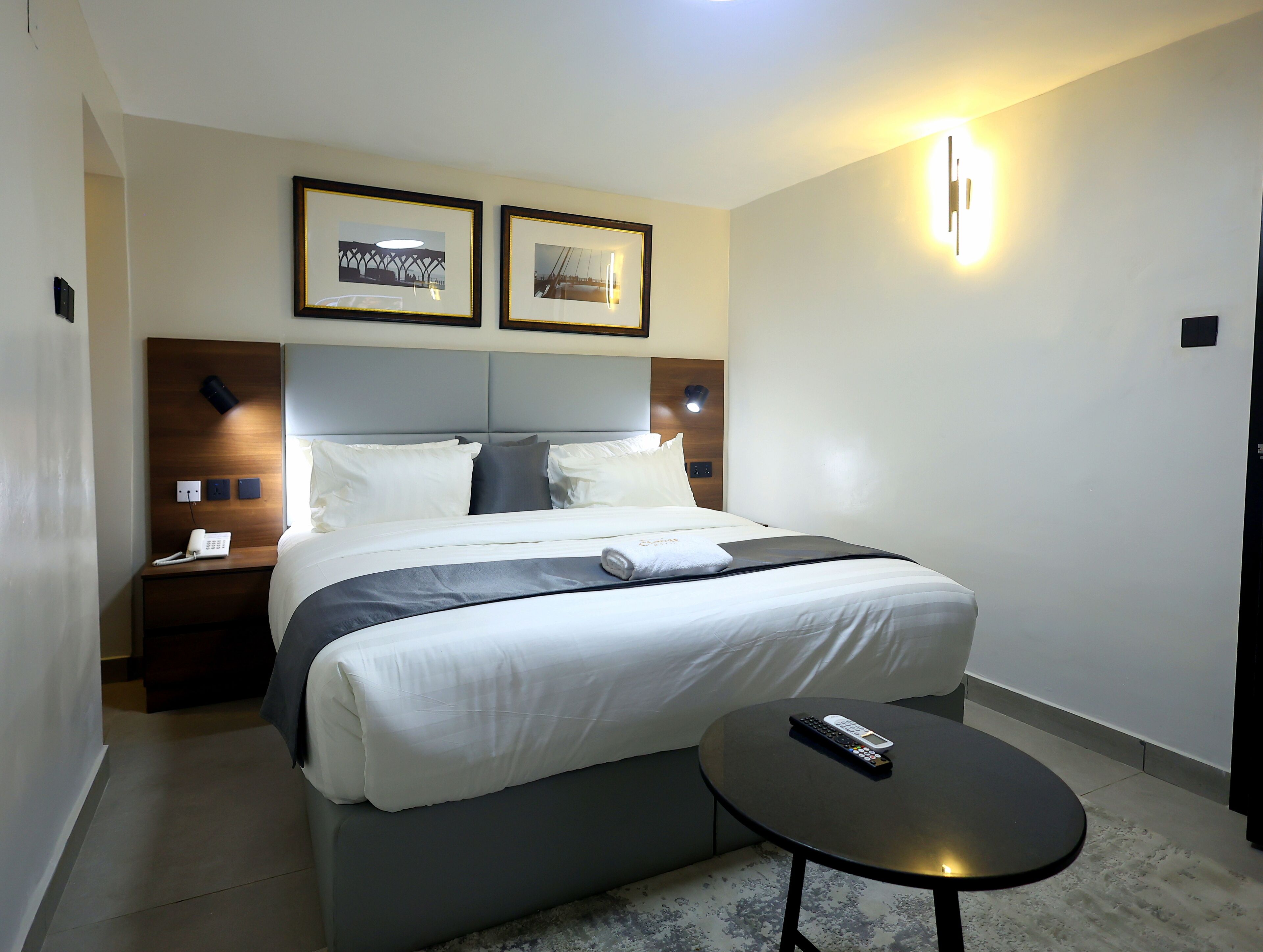 A modern hotel room with a neatly made bed, a bedside table with a remote control, and two framed pictures on the wall.