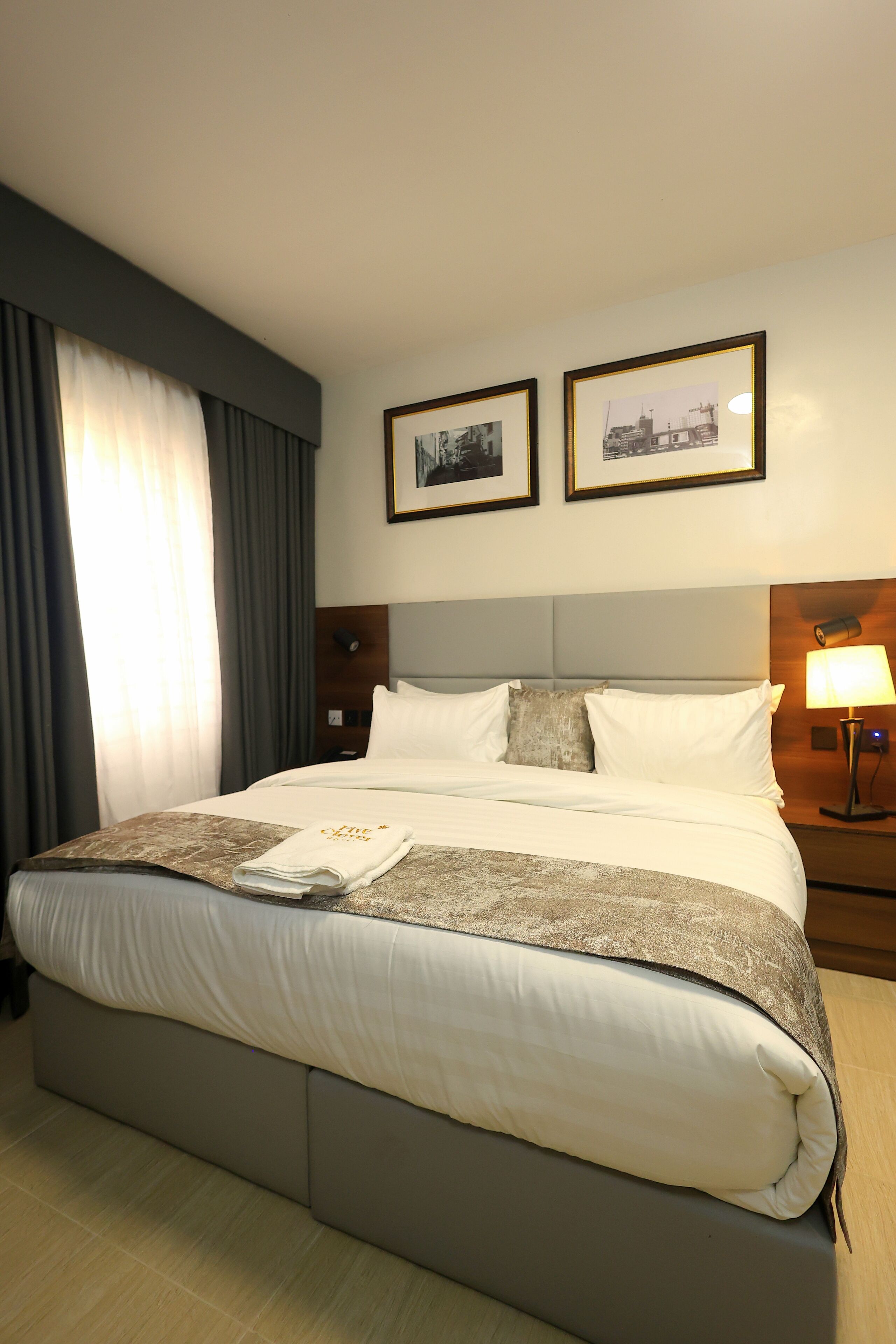 Una habitación de hotel con una cama grande, una mesita de noche con lámpara y dos cuadros enmarcados en la pared.