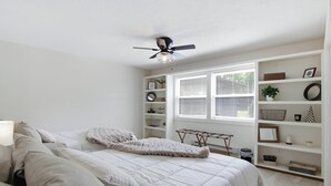 Un dormitorio con una cama, un ventilador de techo, una ventana con persianas y una estantería.