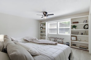 Un dormitorio con una cama, un ventilador de techo, una ventana con persianas y una estantería.