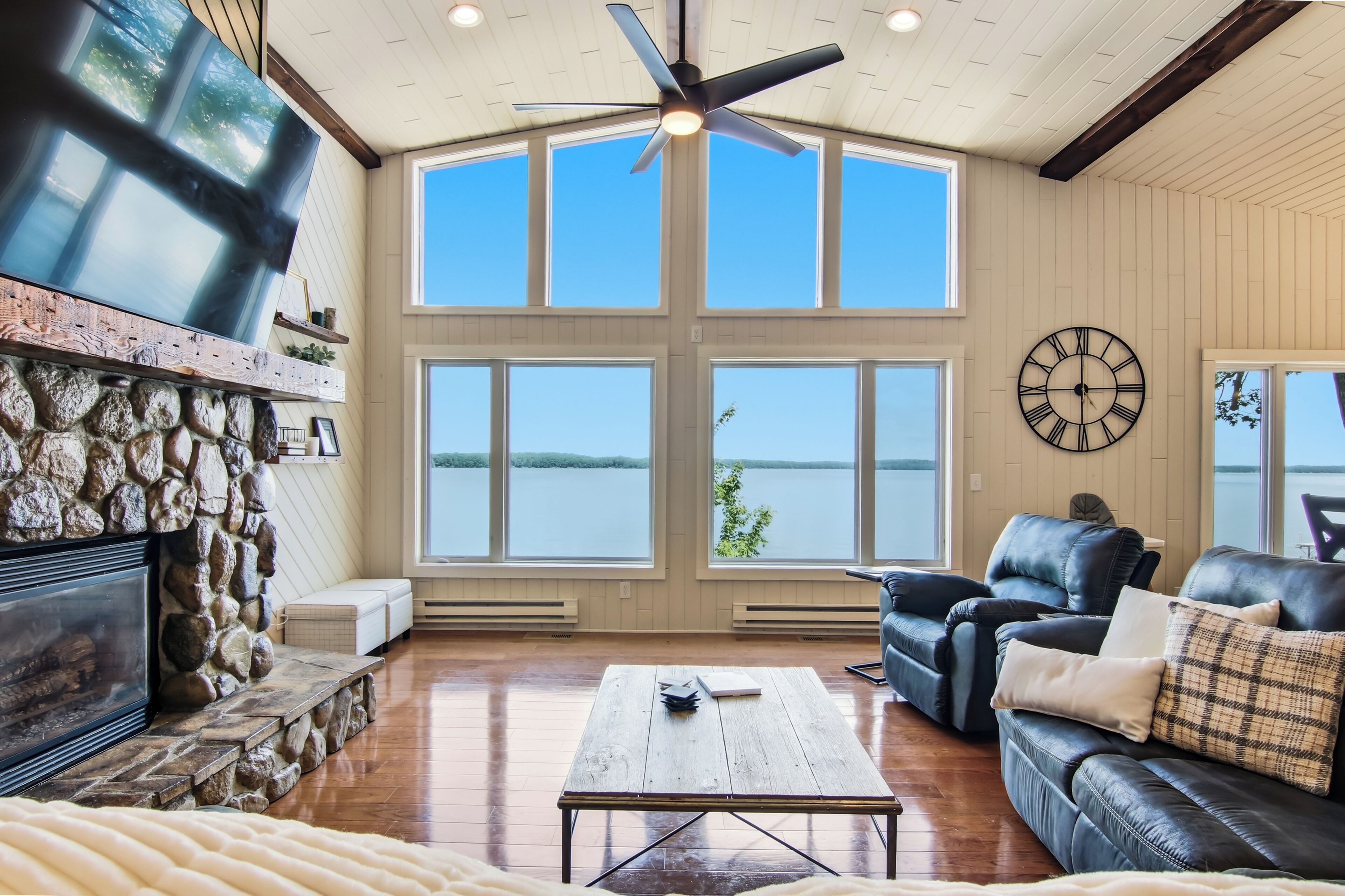 A spacious living room with a stone fireplace, a ceiling fan, large windows overlooking a body of water, and a leather sofa.