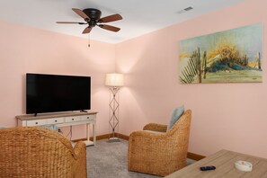 A living room with a flat-screen TV, wicker chairs, a lamp, and a ceiling fan. - Shore Beats Work: Oceanfront Views, Pool, Hot Tub, Sauna, Discounted Golf Rates (Nags Head)