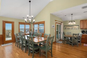 A spacious dining area with a large wooden table and matching chairs, large windows with views, and a kitchen area with wooden cabinets. - Shore Beats Work: Oceanfront Views, Pool, Hot Tub, Sauna, Discounted Golf Rates (Nags Head)
