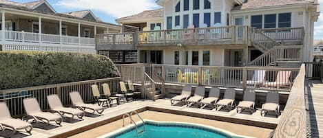 Un área de piscina con sillones reclinables, una terraza de madera y un edificio de varios niveles con balcones.