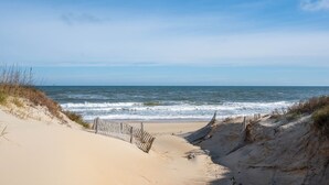 Una playa con una cerca de madera que conduce al agua, dunas arenosas y un cielo despejado.