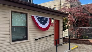 A building with a red door, a window with blinds, and a small American flag decoration. - Charming & luxurious 1-bedroom condo, Kitchen, Washer, dryer, parking, King Bed. (Hagerstown)