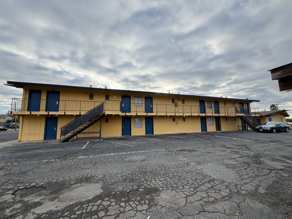 Un motel amarillo con varias habitaciones, un estacionamiento y una escalera que conduce a los pisos superiores.